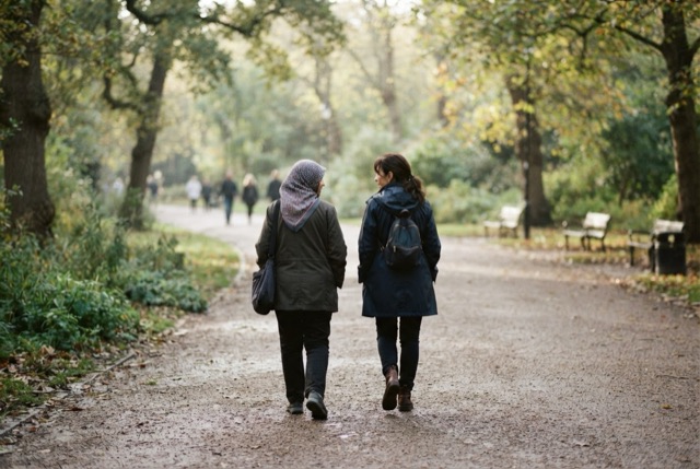 Two women walk side by side along a tree-lined park path